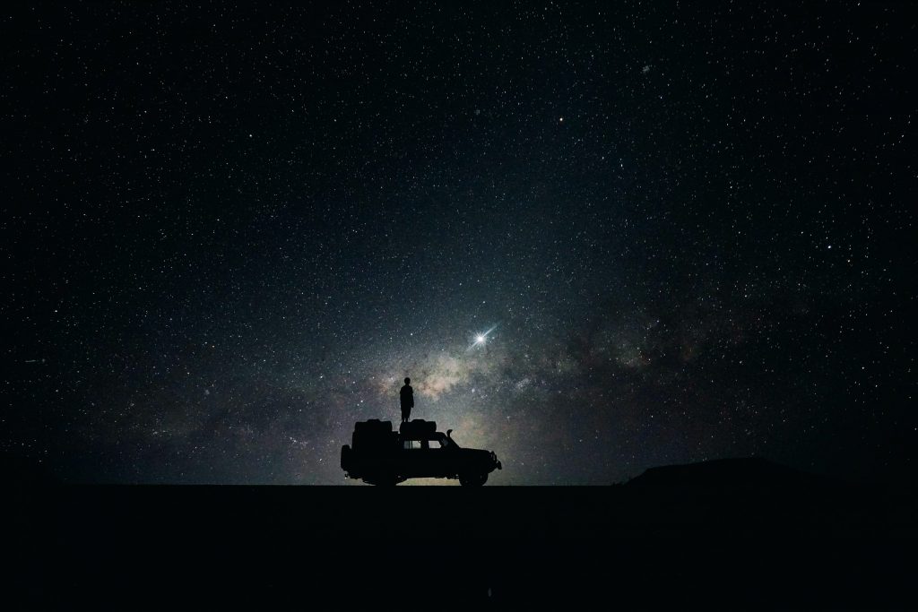 night time shot of a starry sky with the silhouette of a man standing on the top of an SUV in the foreground