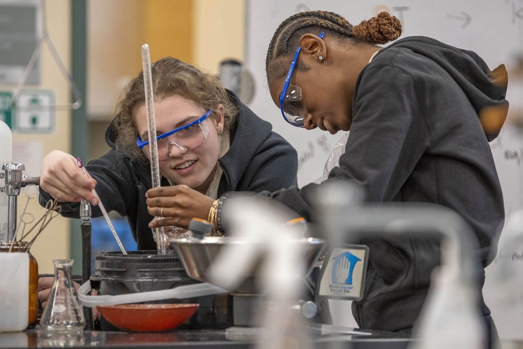 Two students wearing goggles conduct an experiment during a chemistry lab class.