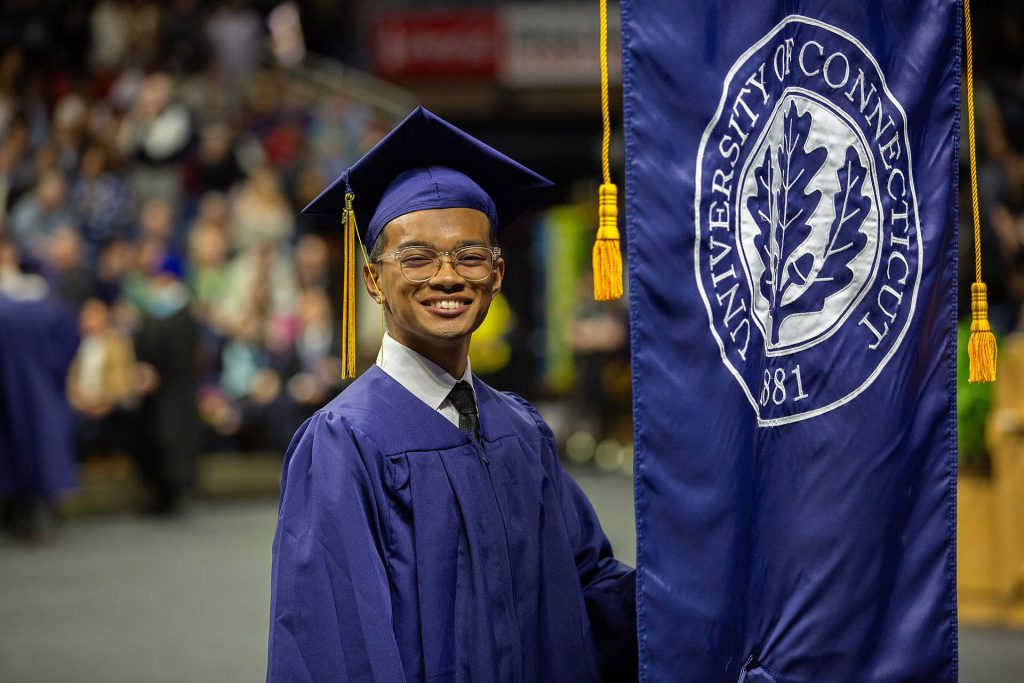 A graduating student in a cap and gown smiles while holding a UConn banner at commencement.