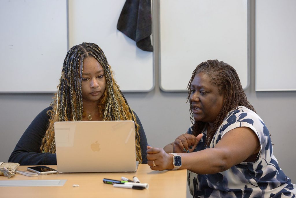 A professor gestures at a laptop screen while meeting with a student during a class.