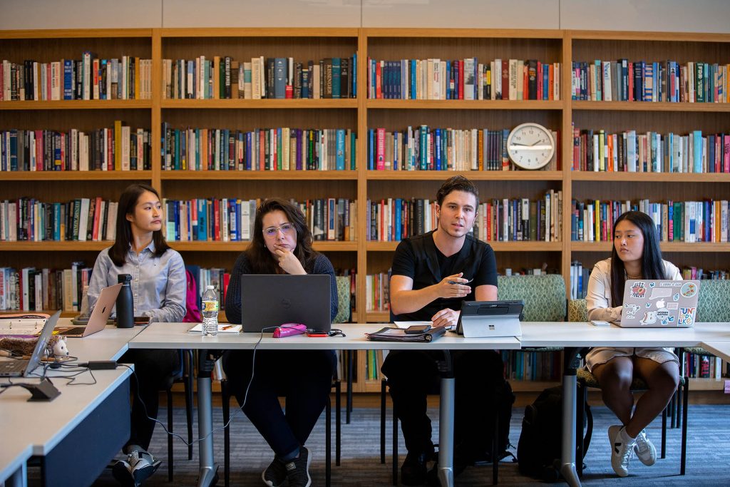 A graduate student gestures while answering a question in a seminar class, while surrounding students look attentively at the instructor.